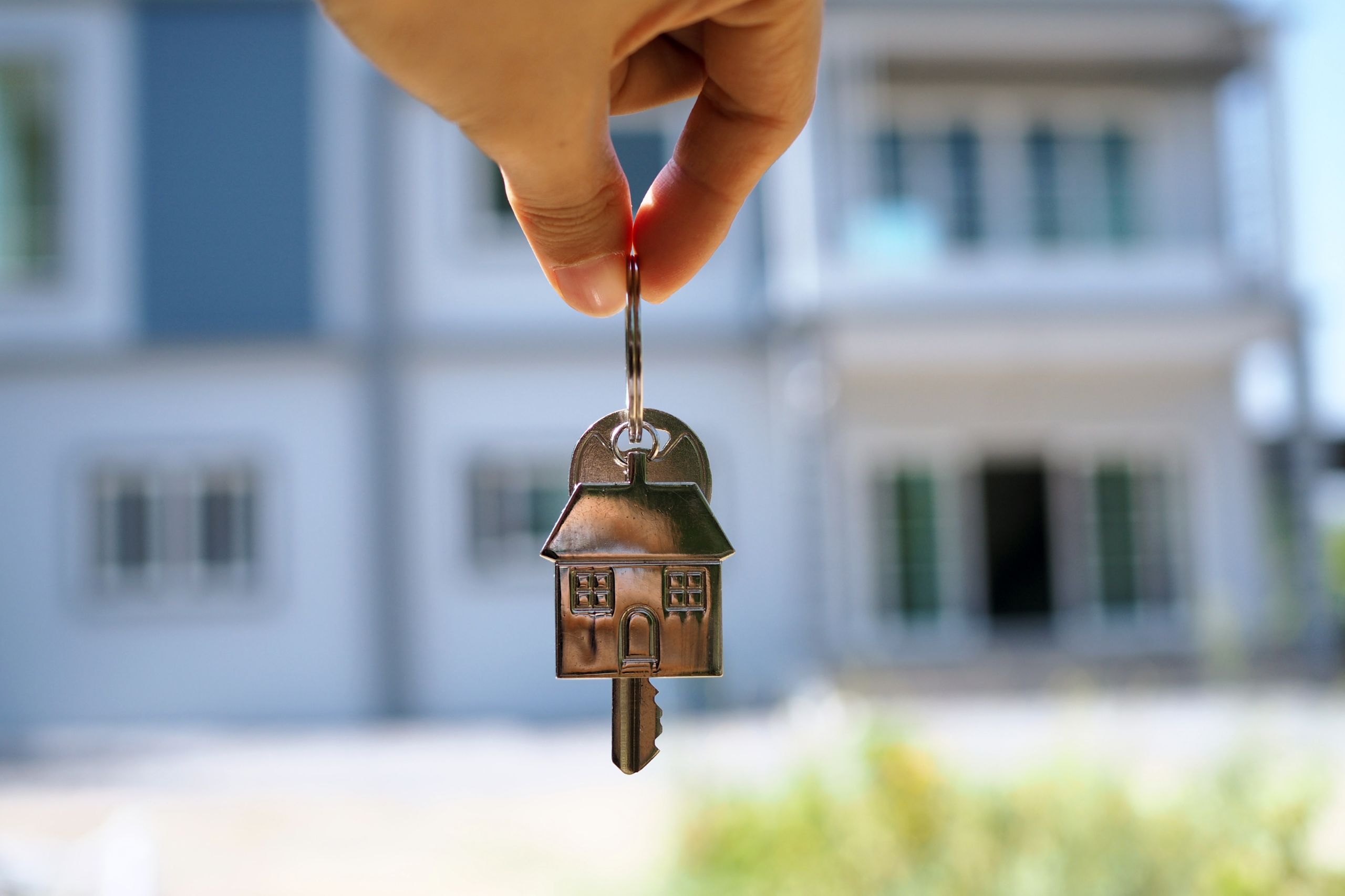 A hand holding a house-shaped key in front of a blurred background of a house, symbolizing home ownership or real estate.