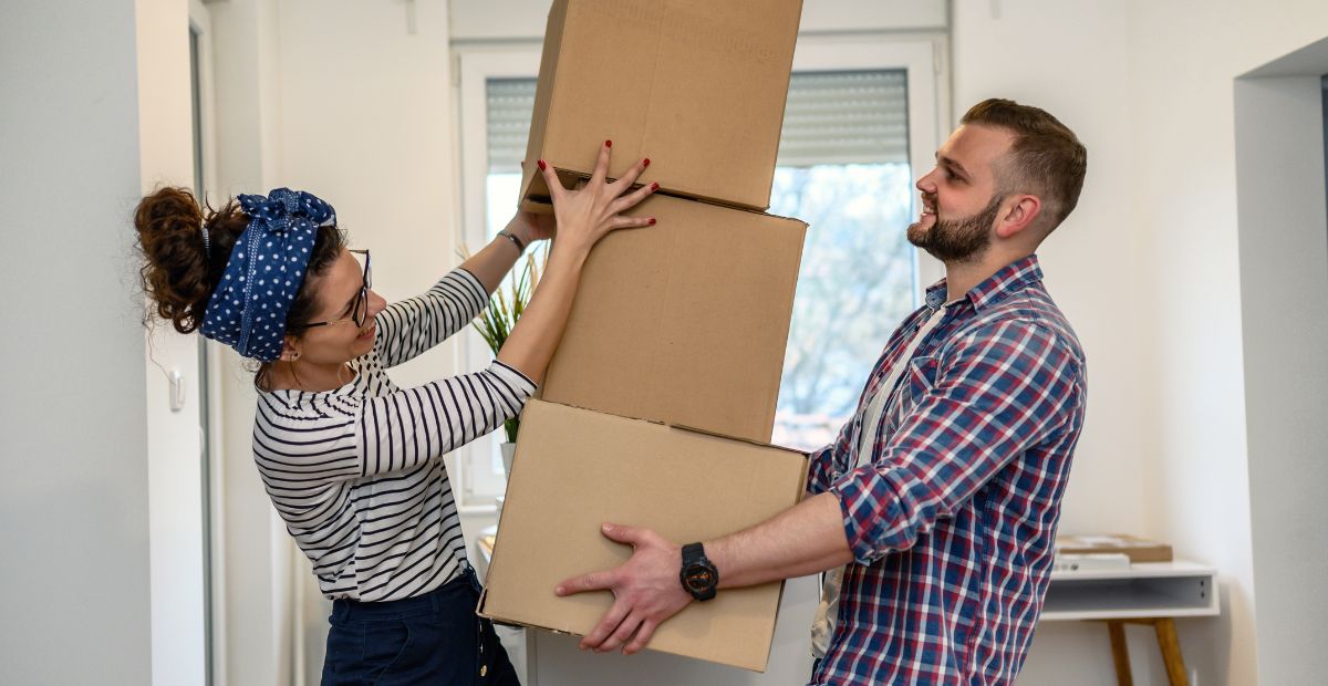 14 Cheerful couple working together to carry a stack of cardboard moving boxes inside their new home.