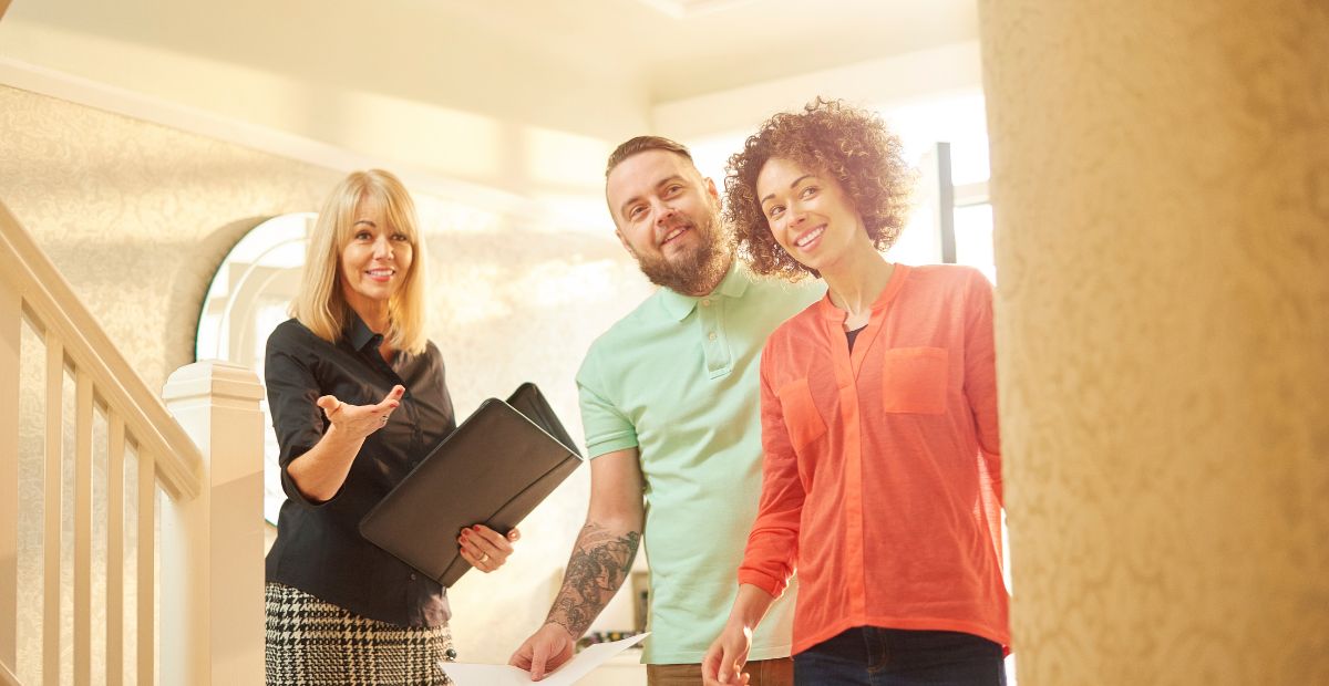 15 Smiling couple being shown a home interior by a real estate agent holding a folder, standing near a staircase.