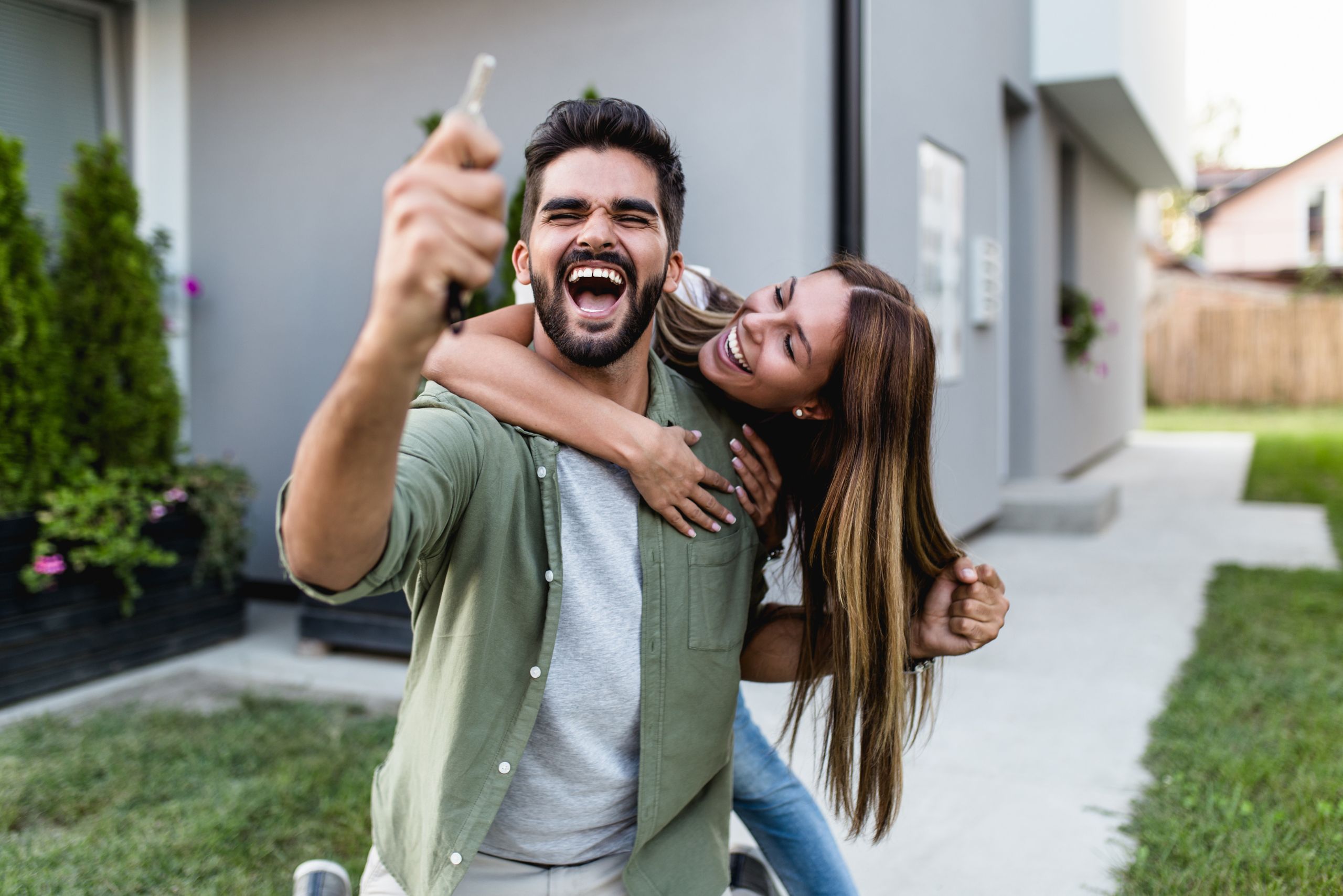 A joyful couple outside their new home, with the man holding up a key and the woman hugging him from behind, celebrating home purchase.