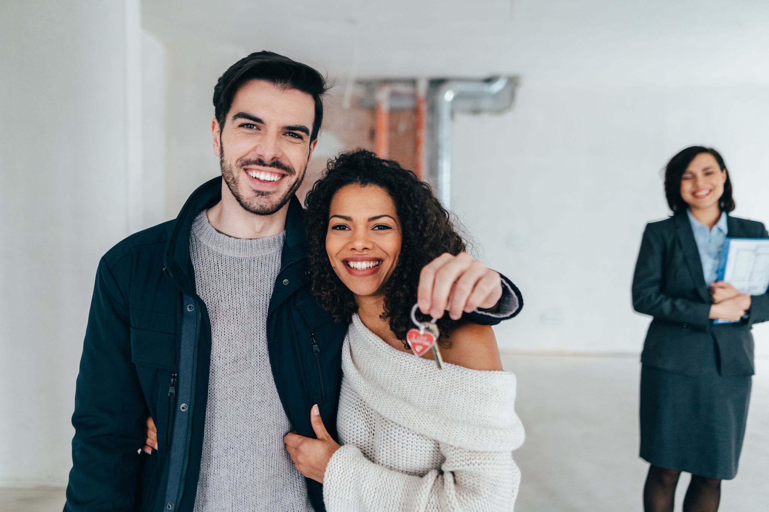 A smiling couple holding up house keys inside a home, with a real estate agent standing in the background, representing a successful home buying experience.